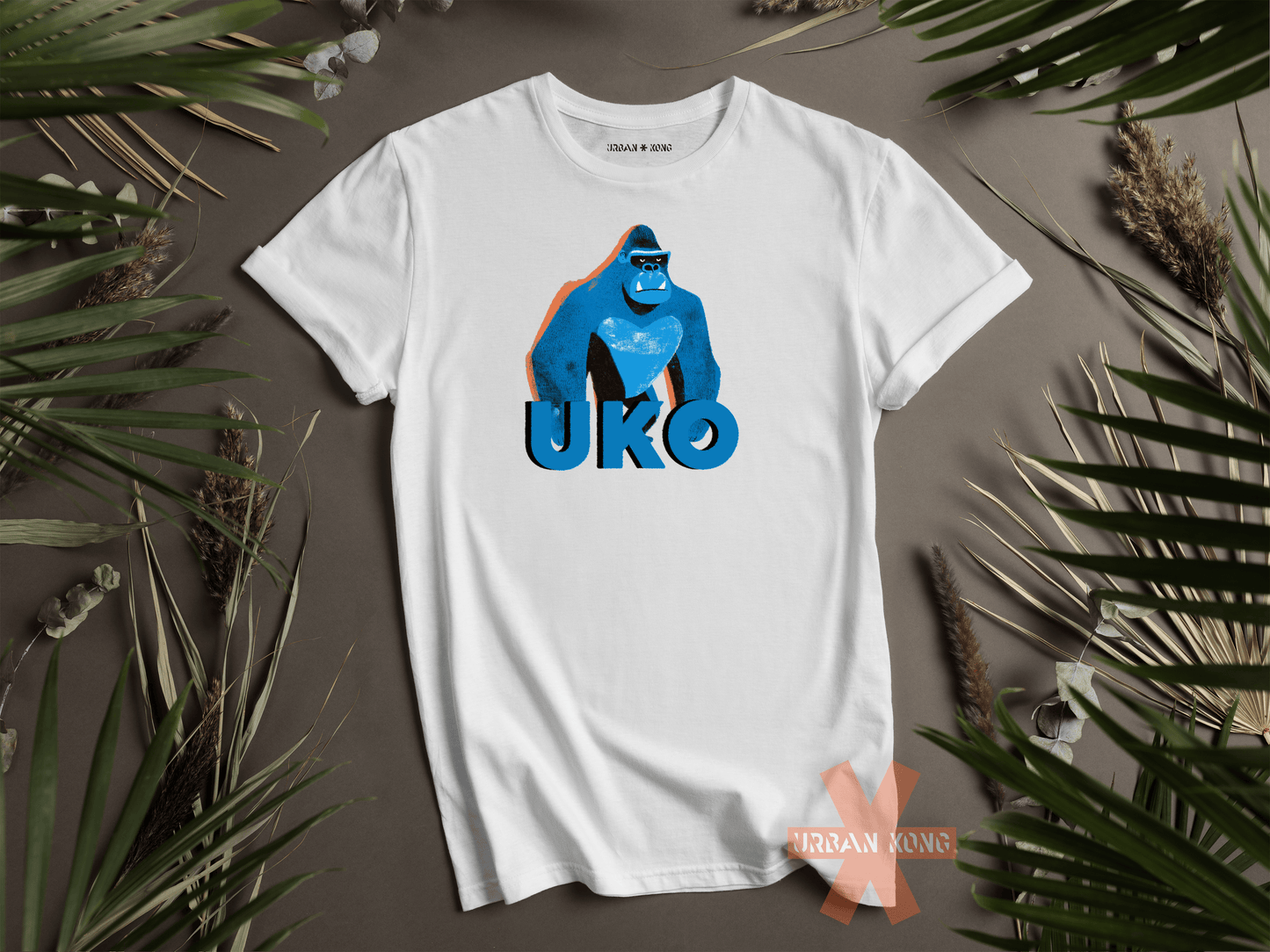 White t-shirt with blue gorilla graphic and 'UKO' text on a gray surface with green leaves.